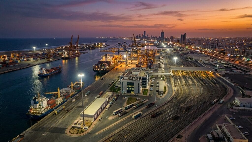 صورة سينمائية لميناء تجاري عند الغروب توضح السفن وحاويات الشحن وأضواء المدينة الذهبية
